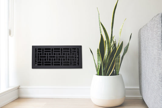 A potted snake plant sits next to a gray sofa and the Madelyn Carter Cast Aluminum Arts & Crafts Vent Cover in black, mounted on a light wall in a bright, modern room.