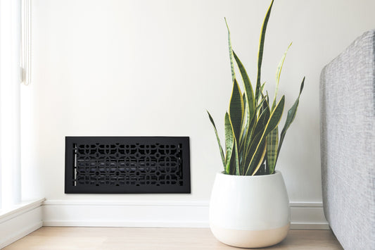 A potted snake plant sits on a light wood floor next to a gray couch and a Madelyn Carter Cast Aluminum Cathedral Vent Cover in black on a white wall.