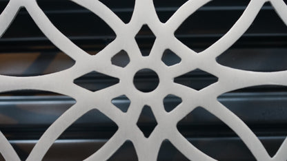 Close-up of the Madelyn Carter Cast Brass Empire Vent Cover in Brushed Nickel, featuring geometric curves and a central hole, set against a dark, blurred background with horizontal lines.