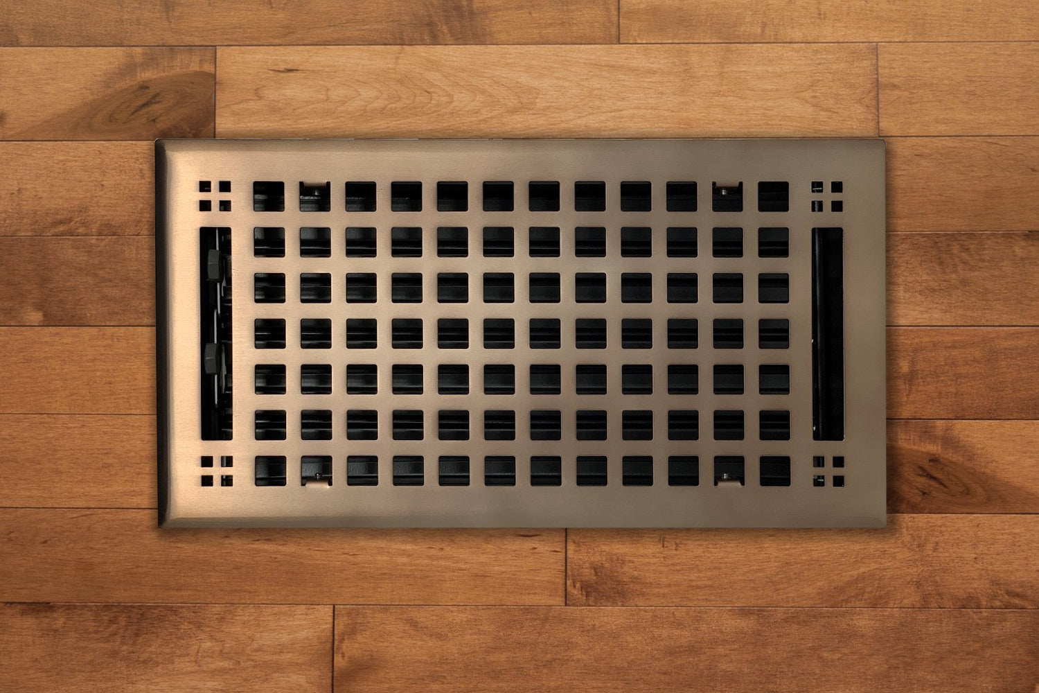 The Madelyn Carter Steel Artisan Vent Covers in oil rubbed bronze feature a grid pattern and are seamlessly installed in light brown wooden flooring, combining durability with elegant style.