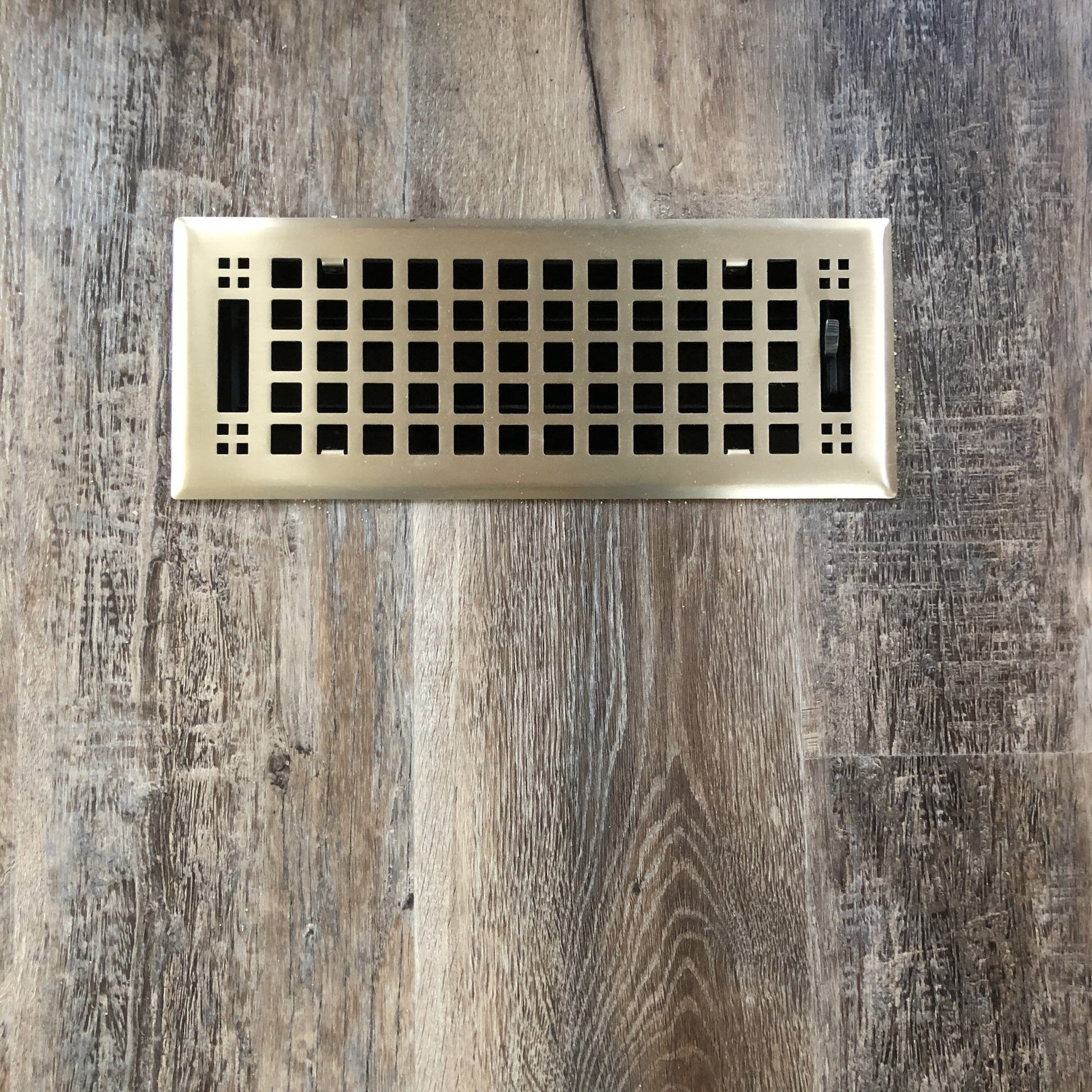 A Madelyn Carter Steel Artisan Vent Cover in brushed brass with a grid pattern is installed on a wood-patterned floor, but the product description only includes CSS code, not details about the actual vent cover.