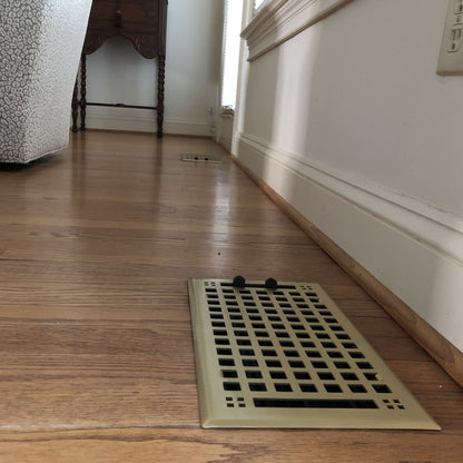 The Madelyn Carter Steel Artisan Vent Covers in brushed brass enhance wooden floors, with two small black objects atop one vent and another cover seen near the wall, adding elegant style to any interior.