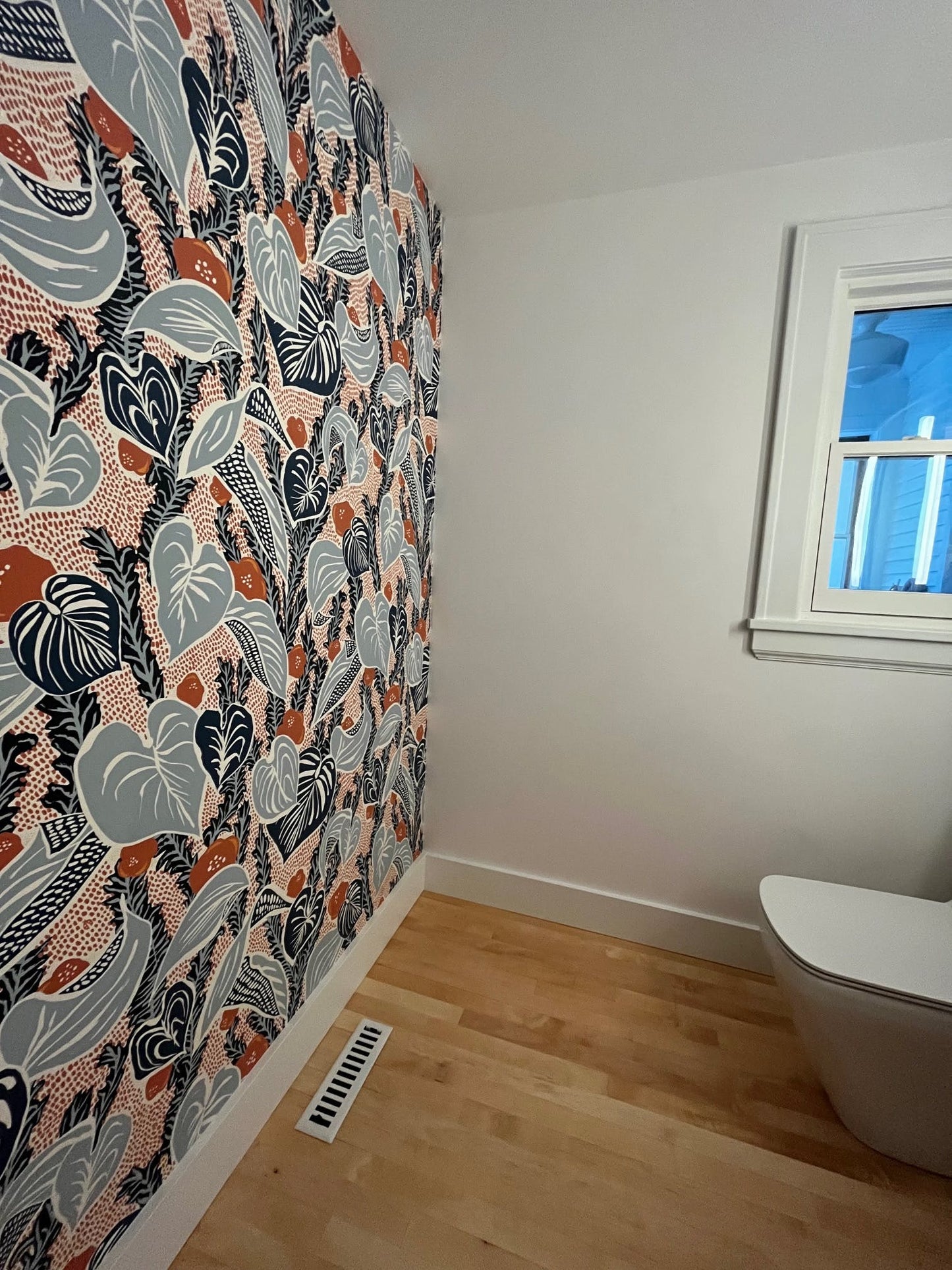 A room with a colorful patterned wallpapered wall, Madelyn Carter Steel Modern Chic Vent Covers in white on the wooden floor, a window, and part of a white toilet.