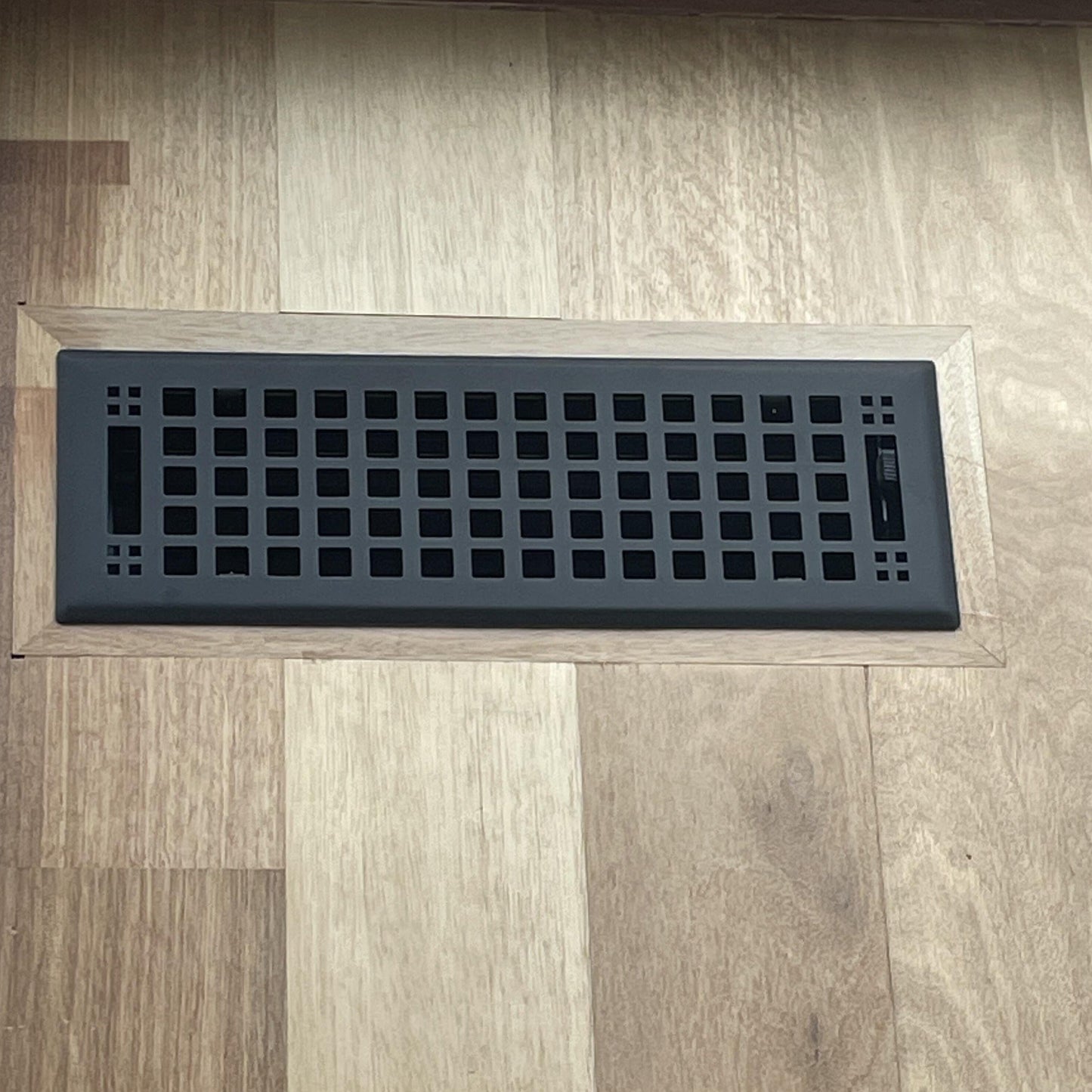 The Madelyn Carter Steel Artisan Vent Cover in black, featuring a grid pattern, is installed in a light wood floor with varied shades; only CSS code and HTML structure are shown, with no product description text provided.