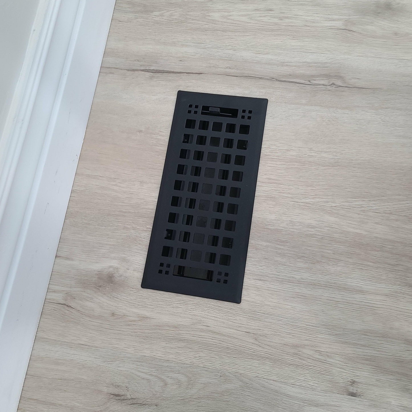 A Madelyn Carter Steel Artisan Vent Cover in black, featuring a rectangular grid design, is installed in light wood flooring near a white baseboard—displayed using only CSS code and HTML structure, with no actual product description text.