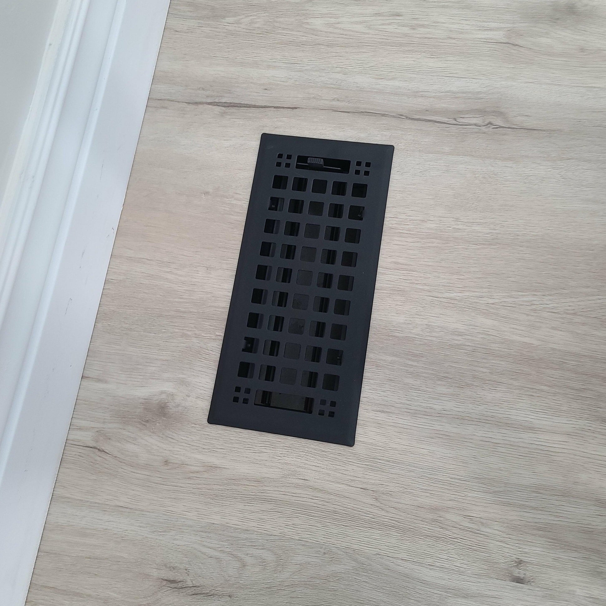 A Madelyn Carter Steel Artisan Vent Cover in black, featuring a rectangular grid design, is installed in light wood flooring near a white baseboard—displayed using only CSS code and HTML structure, with no actual product description text.