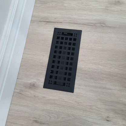 A Madelyn Carter Steel Artisan Vent Cover in black, featuring a rectangular grid design, is installed in light wood flooring near a white baseboard—displayed using only CSS code and HTML structure, with no actual product description text.