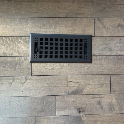 A Madelyn Carter Steel Artisan Vent Cover in black is installed on light brown wood flooring with visible grain; no product description is provided—only CSS code and HTML structure are shown.