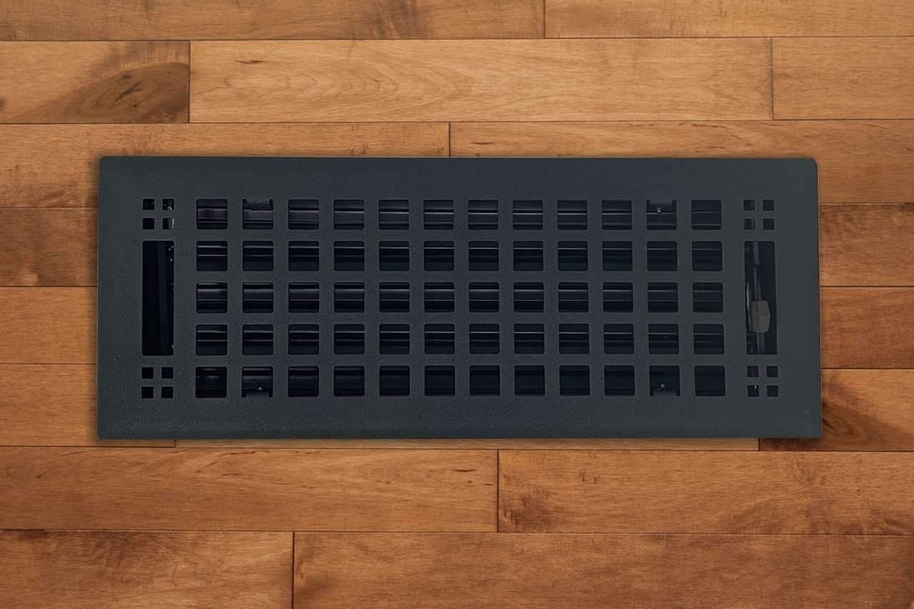 The Madelyn Carter Steel Artisan Vent Cover in black, featuring a sleek grid pattern, is installed in medium brown wood flooring—showcasing stylish design where function typically rules.