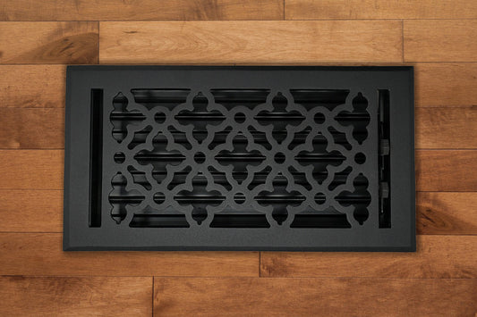 The Madelyn Carter Cast Aluminum Gothic Vent Cover in black is installed on a light brown wooden floor, bringing stylish geometric design and functionality to your space.