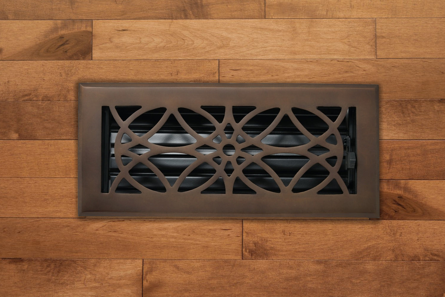 The Madelyn Carter Cast Brass Empire Vent Cover in Oil Rubbed Bronze is installed in light brown hardwood flooring, adding elegance with its geometric pattern.