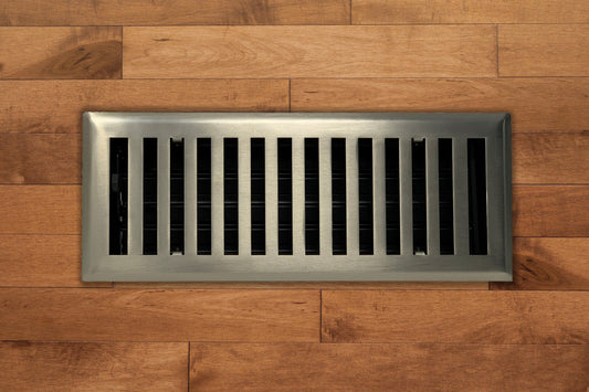 A Madelyn Carter Steel Modern Chic Vent Cover in brushed nickel with black slats is installed into light brown wooden flooring.