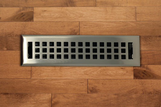 The Madelyn Carter Steel Artisan Vent Cover in Brushed Nickel features a grid pattern, installed in hardwood flooring to provide a stylish and durable finish.