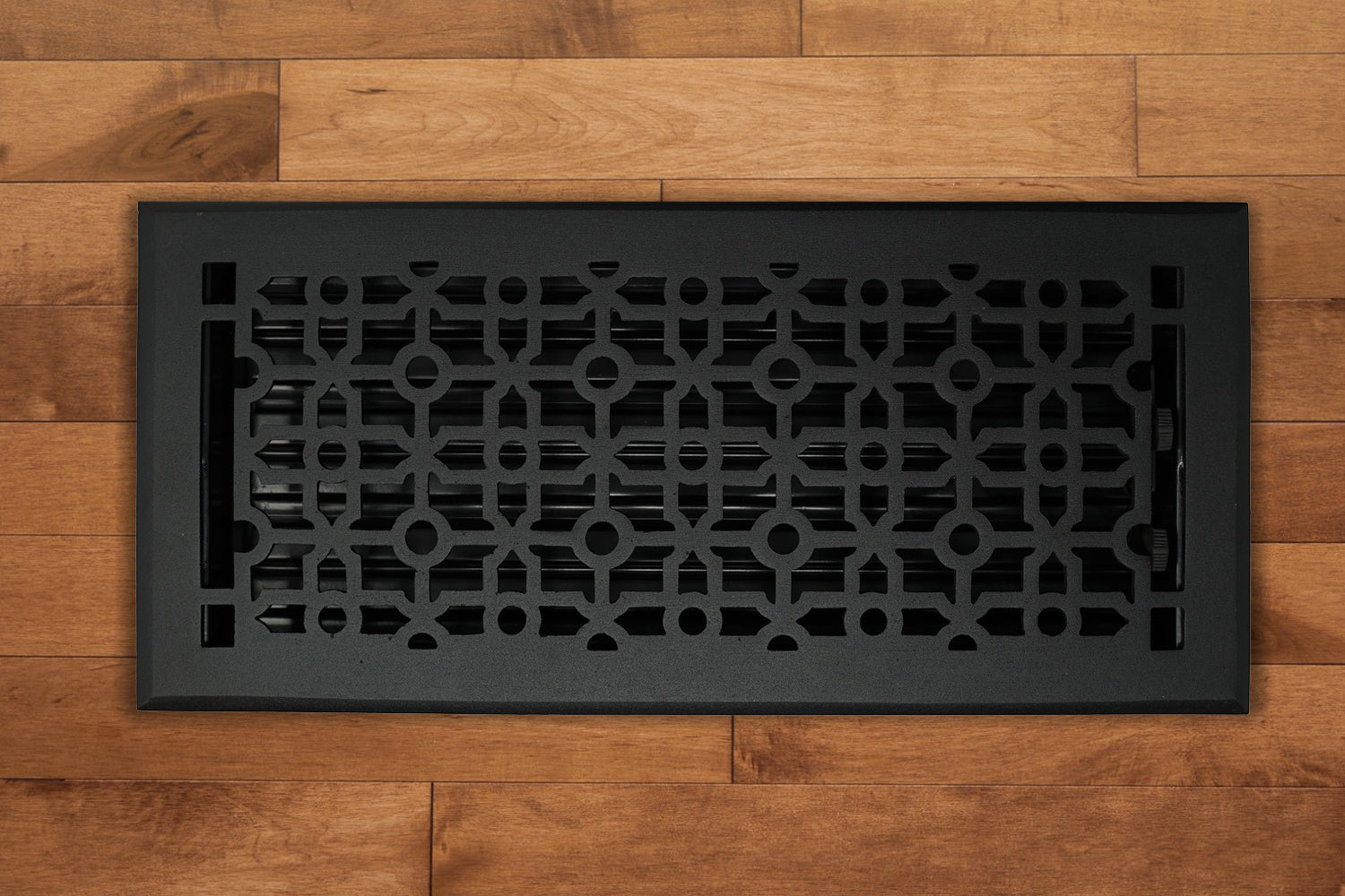 The Madelyn Carter Cast Aluminum Cathedral Vent Cover in black, featuring a geometric pattern, is displayed on a light brown wooden floor.