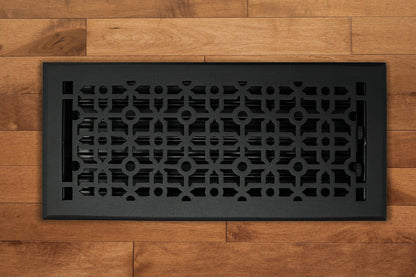 The Madelyn Carter Cast Aluminum Cathedral Vent Cover in black, featuring a geometric pattern, is displayed on a light brown wooden floor.