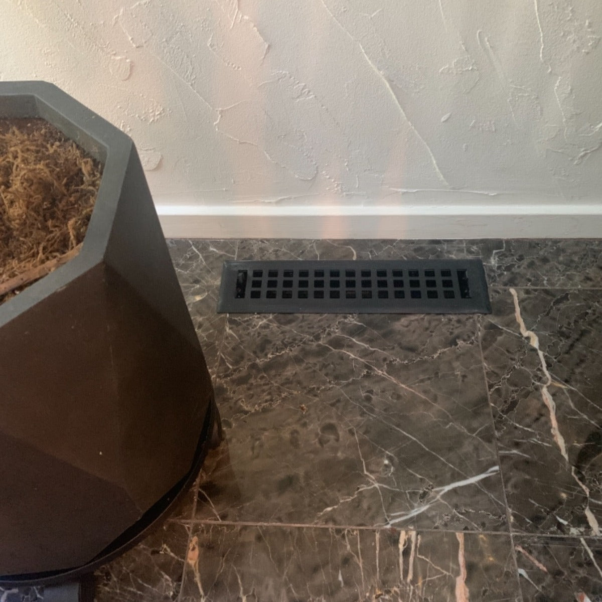 A Madelyn Carter Steel Artisan Vent Cover in black is installed in dark marble tile near a textured white wall, with a geometric planter partly visible on the left; only CSS code and HTML structure are shown, not an actual product description.