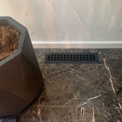 A Madelyn Carter Steel Artisan Vent Cover in black is installed in dark marble tile near a textured white wall, with a geometric planter partly visible on the left; only CSS code and HTML structure are shown, not an actual product description.