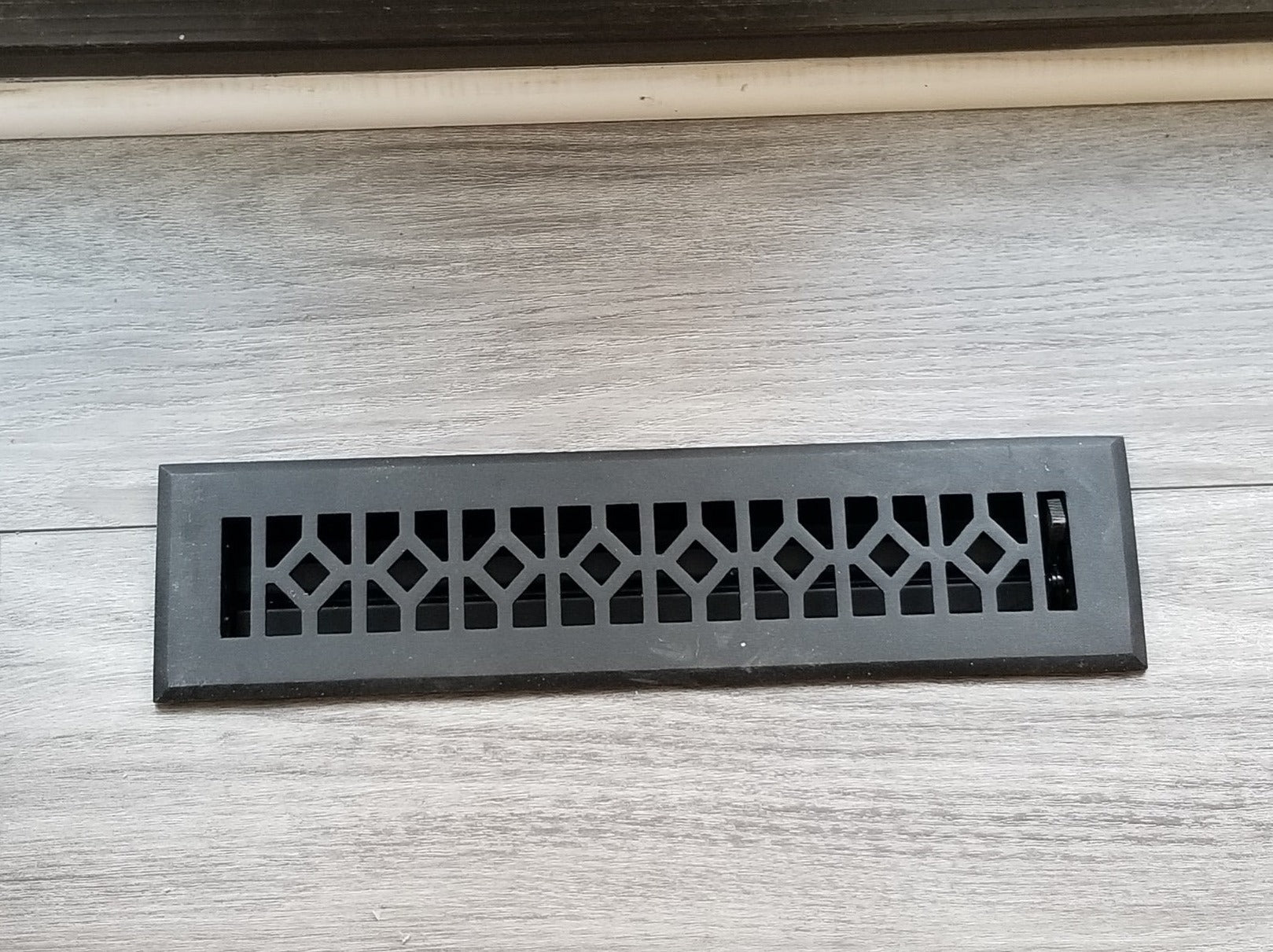 A Madelyn Carter Cast Aluminum Roman Vent Cover in black with a geometric design is installed in a light gray wood floor beside a white baseboard.