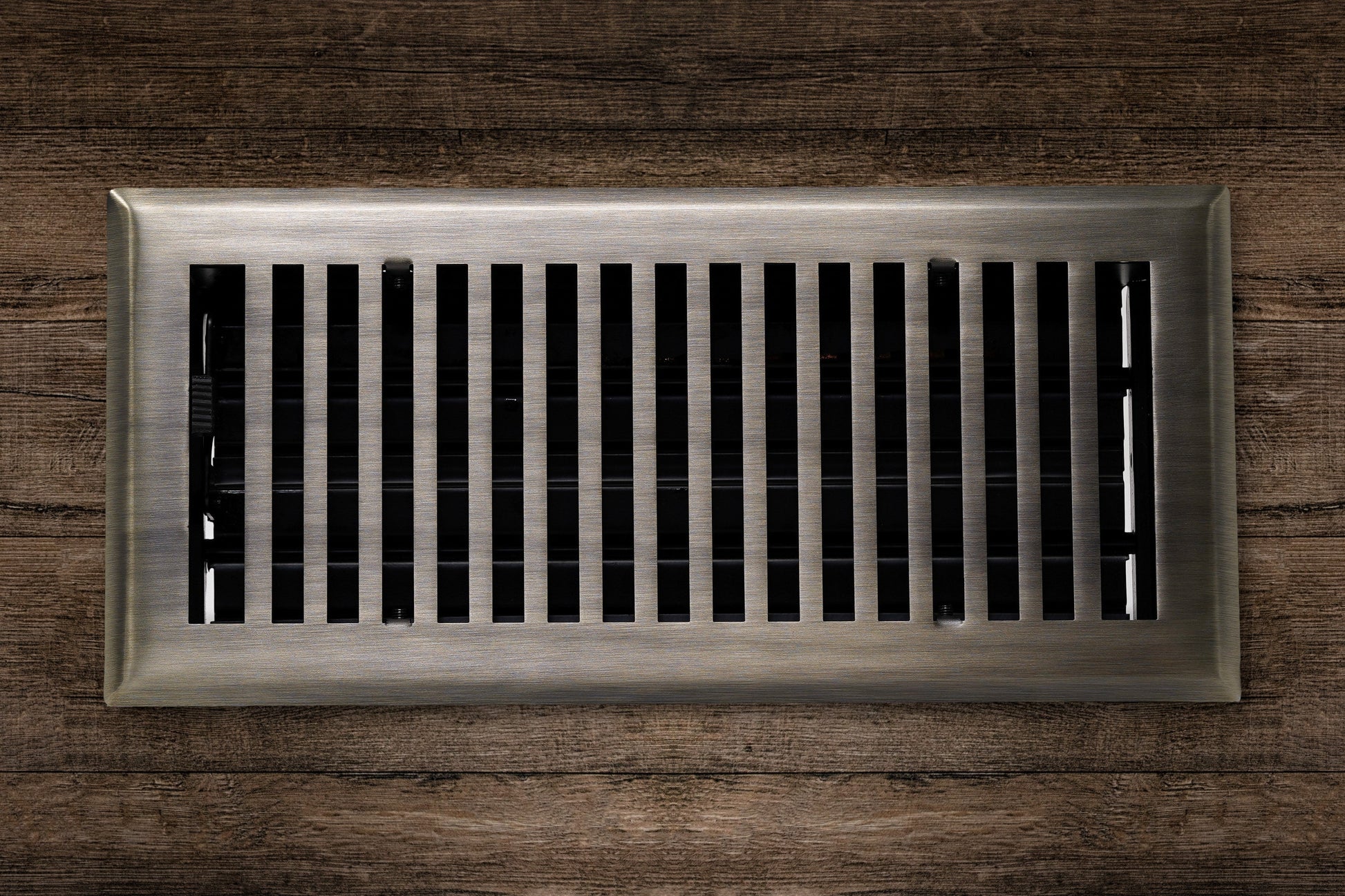 The Madelyn Carter Cast Brass Contemporary Vent Cover in Antique Brass is installed in a wooden surface, providing both functionality and modern style with its vertical metal slats.