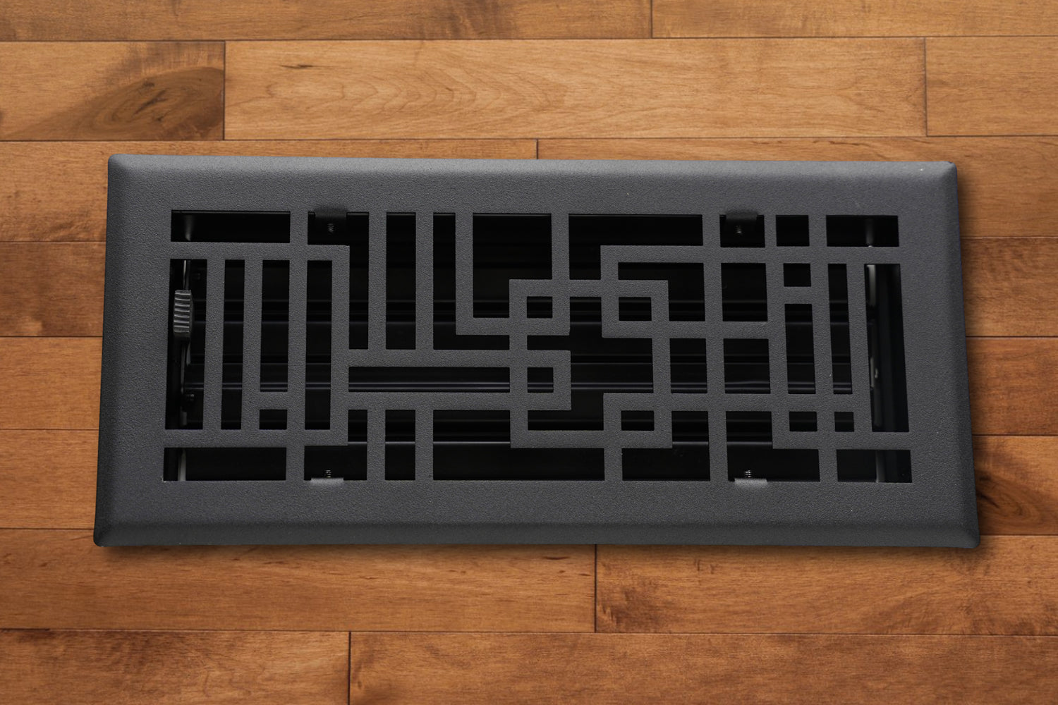 The Madelyn Carter Steel Capitman Art Deco Vent Cover in black, featuring a geometric pattern, is installed in light brown hardwood flooring.