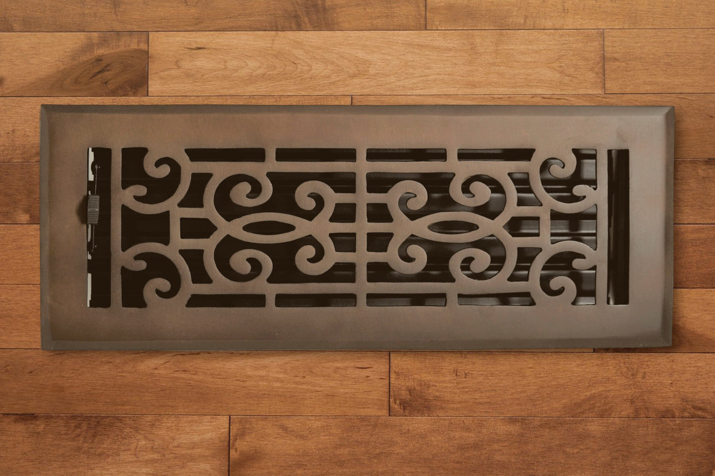 A Madelyn Carter Cast Brass Baroque Vent Cover in oil rubbed bronze with an ornate pattern is set in light brown wooden flooring.