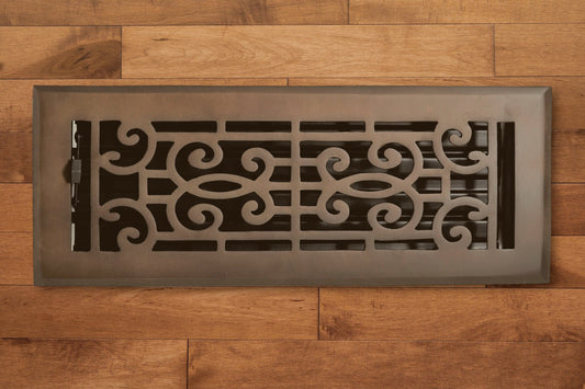 A Madelyn Carter Cast Brass Baroque Vent Cover in oil rubbed bronze with an ornate pattern is set in light brown wooden flooring.
