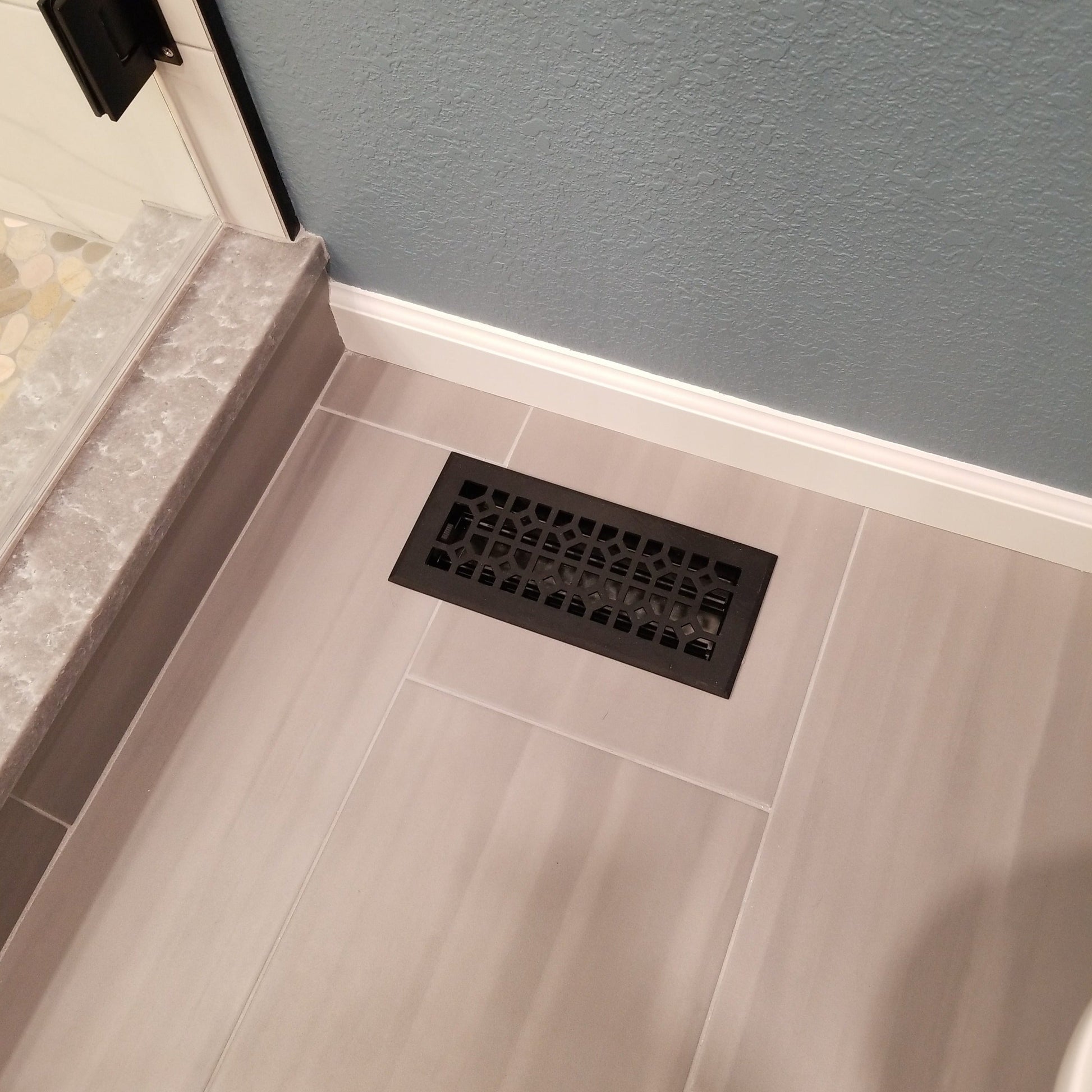 The Madelyn Carter Cast Aluminum Roman Vent Cover in black is set into light gray tile flooring beside a shower with a stone threshold and a blue textured wall.