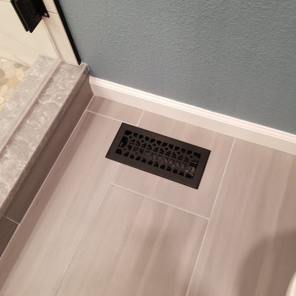 The Madelyn Carter Cast Aluminum Roman Vent Cover in black is set into light gray tile flooring beside a shower with a stone threshold and a blue textured wall.
