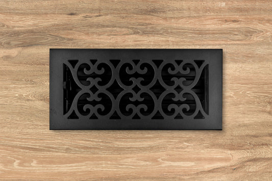 The Madelyn Carter Cast Iron Heirloom Vent Cover in black features a scroll pattern, set into a light wood floor for an elegant decorative accent.