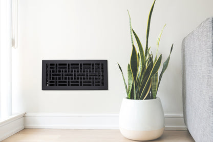 A potted snake plant sits next to a gray sofa and the Madelyn Carter Cast Aluminum Arts & Crafts Vent Cover in black, mounted on a light wall in a bright, modern room.