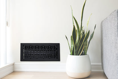 A potted snake plant sits on a light wood floor next to a gray couch and a Madelyn Carter Cast Aluminum Cathedral Vent Cover in black on a white wall.