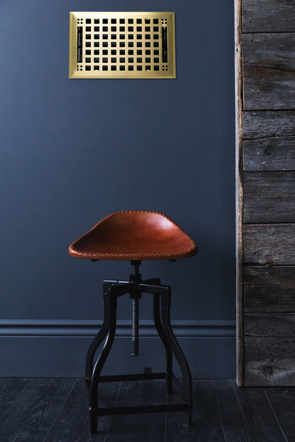 A brown leather industrial-style stool with a sturdy metal base stands against a dark wall. Above, the Steel Artisan Vent Cover by Madelyn Carter in brushed brass adds character, while reclaimed wood paneling to the right enhances the rustic appeal.