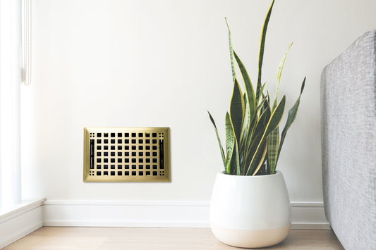 A potted snake plant rests on a light wood floor beside a gray sofa and a Madelyn Carter Steel Artisan Vent Cover in Brushed Brass, adding natural charm and style to your living space.