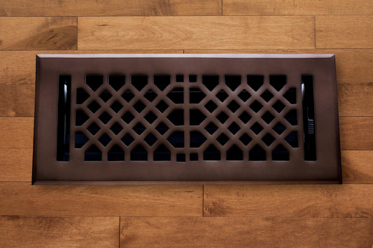The Madelyn Carter Cast Brass Vintage Industrial Vent Cover in oil rubbed bronze features a geometric lattice pattern, adding a striking accent to wooden flooring.