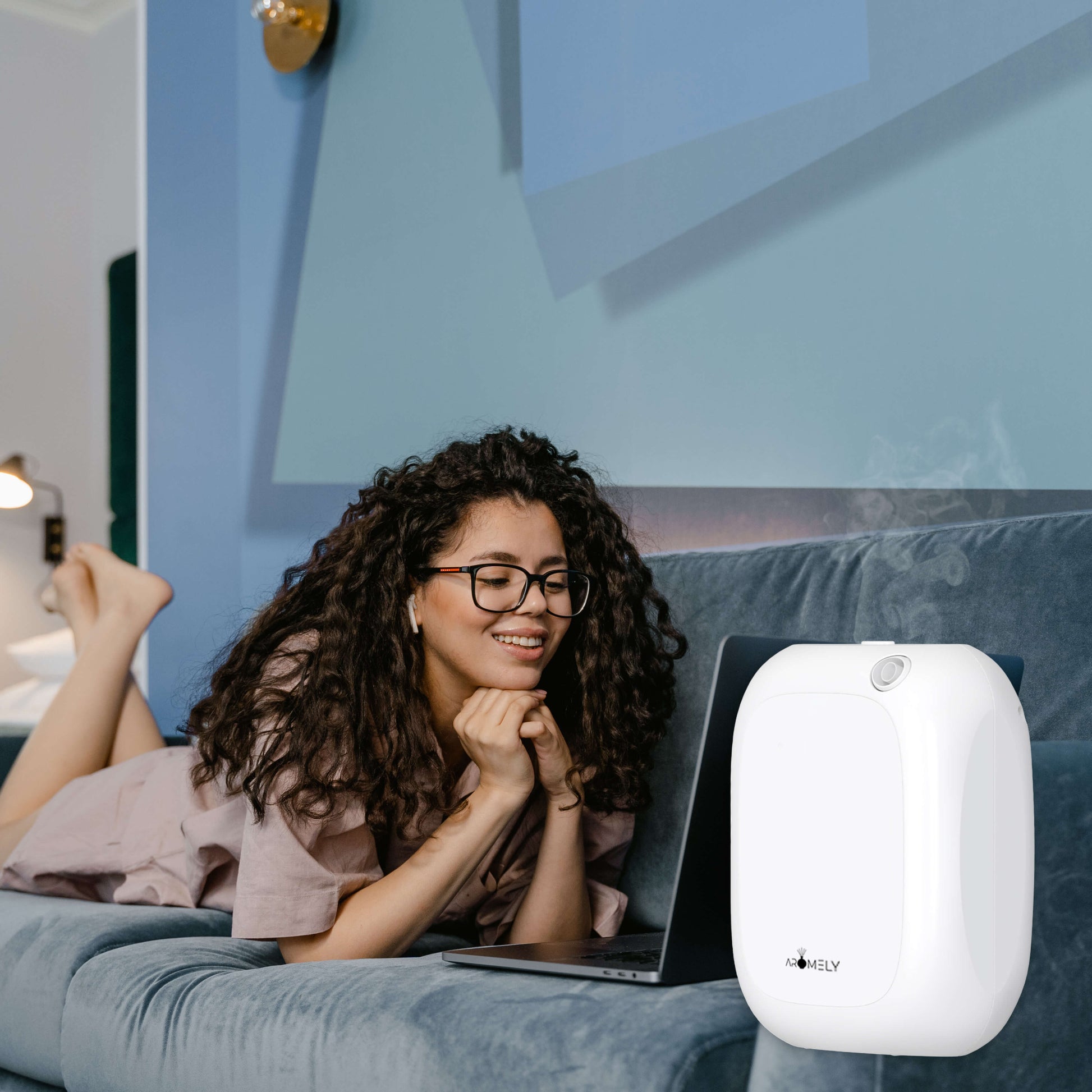 A woman lying on a couch uses a laptop, with the AROMELY Aromely Smart Bluetooth Scent Diffuser for up to 1,500 sq ft nearby, emitting mist.
