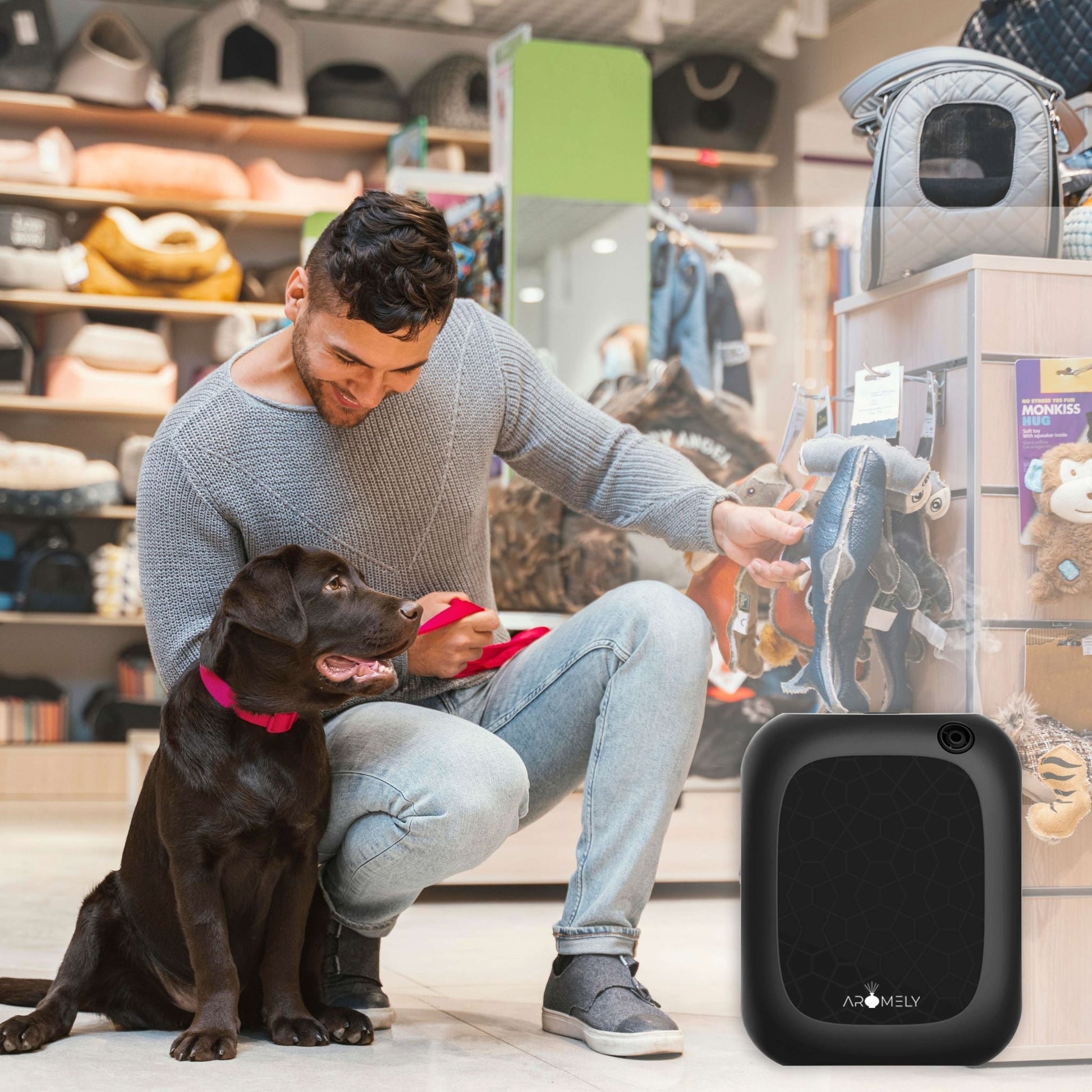 A man kneels beside a brown dog in a pet store, holding a red leash, with various pet products and the AROMELY Smart Bluetooth Scent Diffuser (covers up to 1,500 Sqft., cold diffusion technology) displayed nearby.