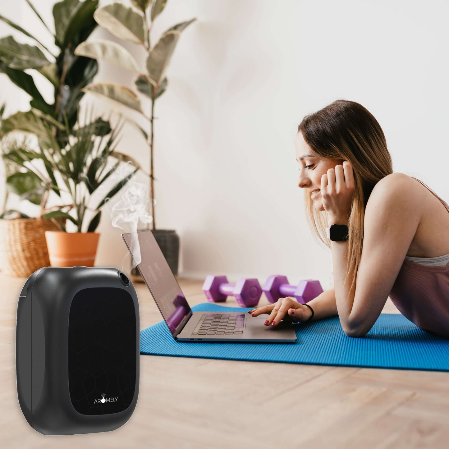 A woman lies on a blue exercise mat using a laptop, with purple dumbbells, an AROMELY Smart Bluetooth Scent Diffuser for up to 1,500 sq ft., and a black air purifier nearby in a bright room with plants.