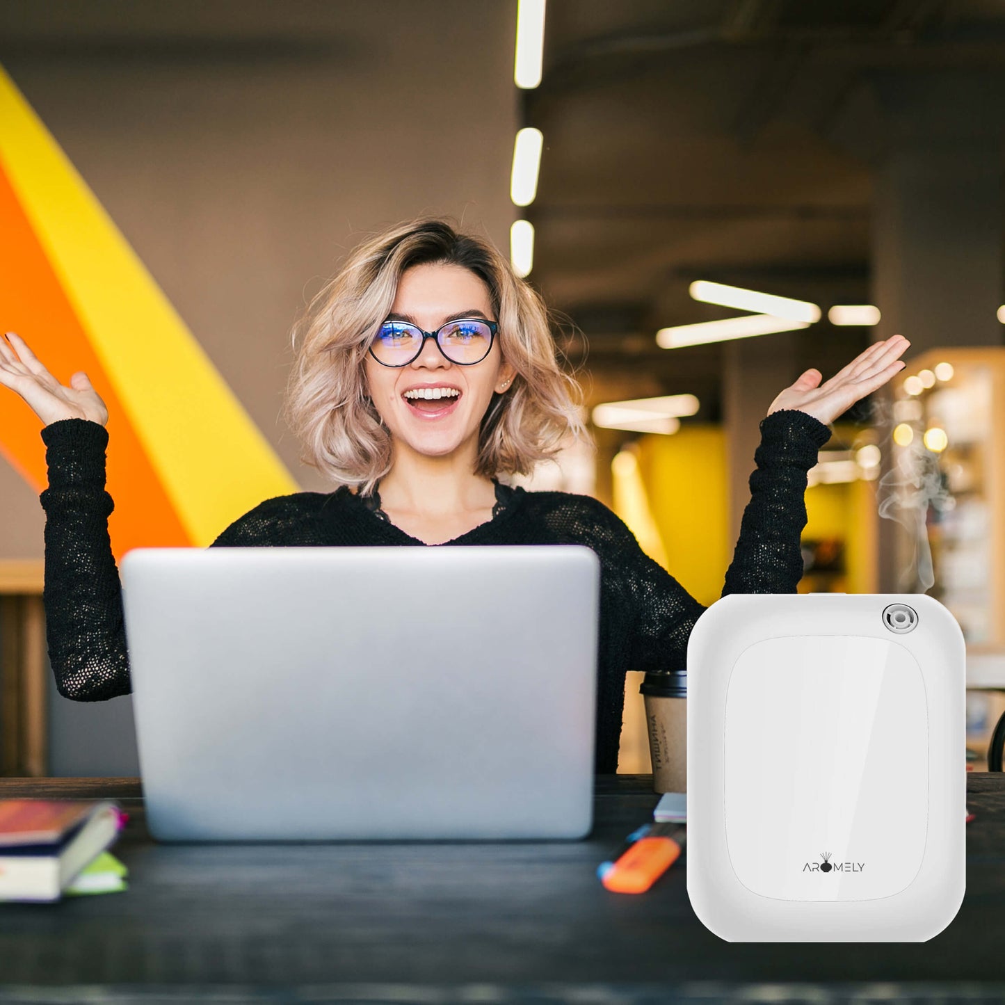 A woman with glasses smiles and raises her hands at a desk with a laptop, books, and the Aromely Smart Bluetooth Scent Diffuser by AROMELY in a modern, colorful workspace.