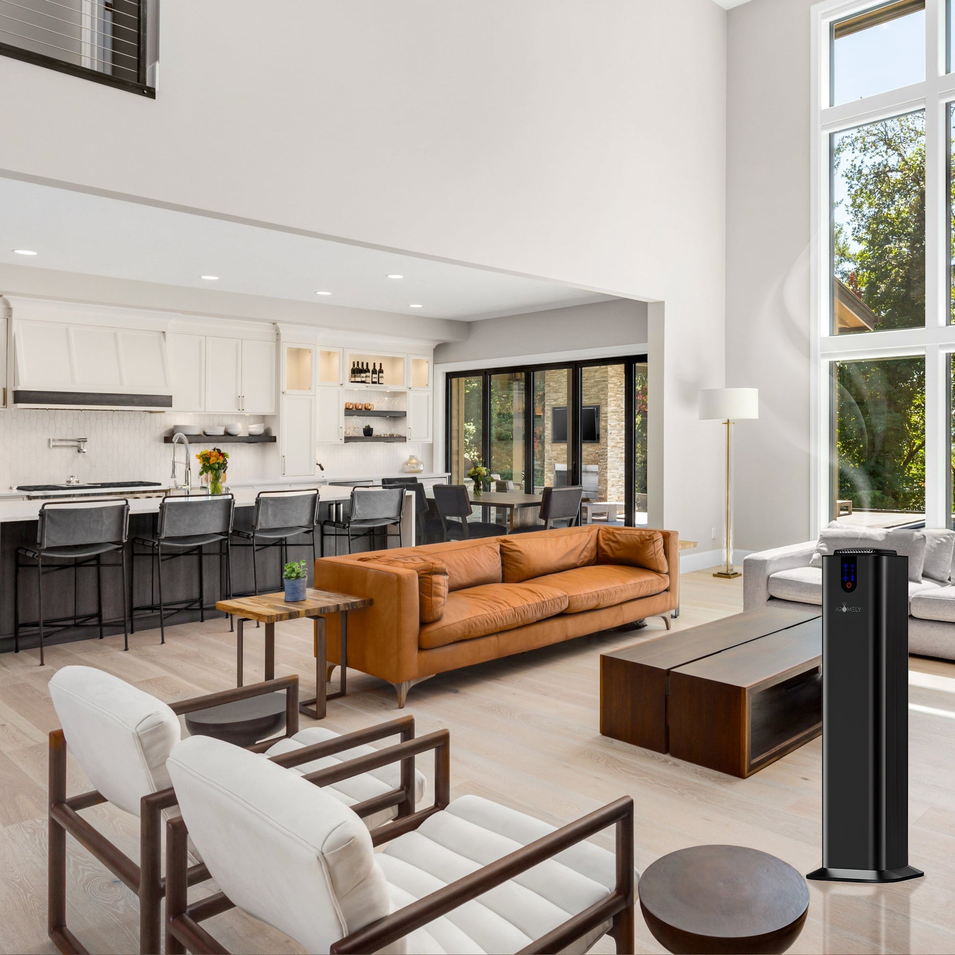 Modern open-concept living room and kitchen featuring a brown leather sofa, white armchairs, bar seating, large windows, a black tower fan, and the AROMELY Aromely Smart Bluetooth Scent Diffuser for up to 2,000 SQSF in the foreground.