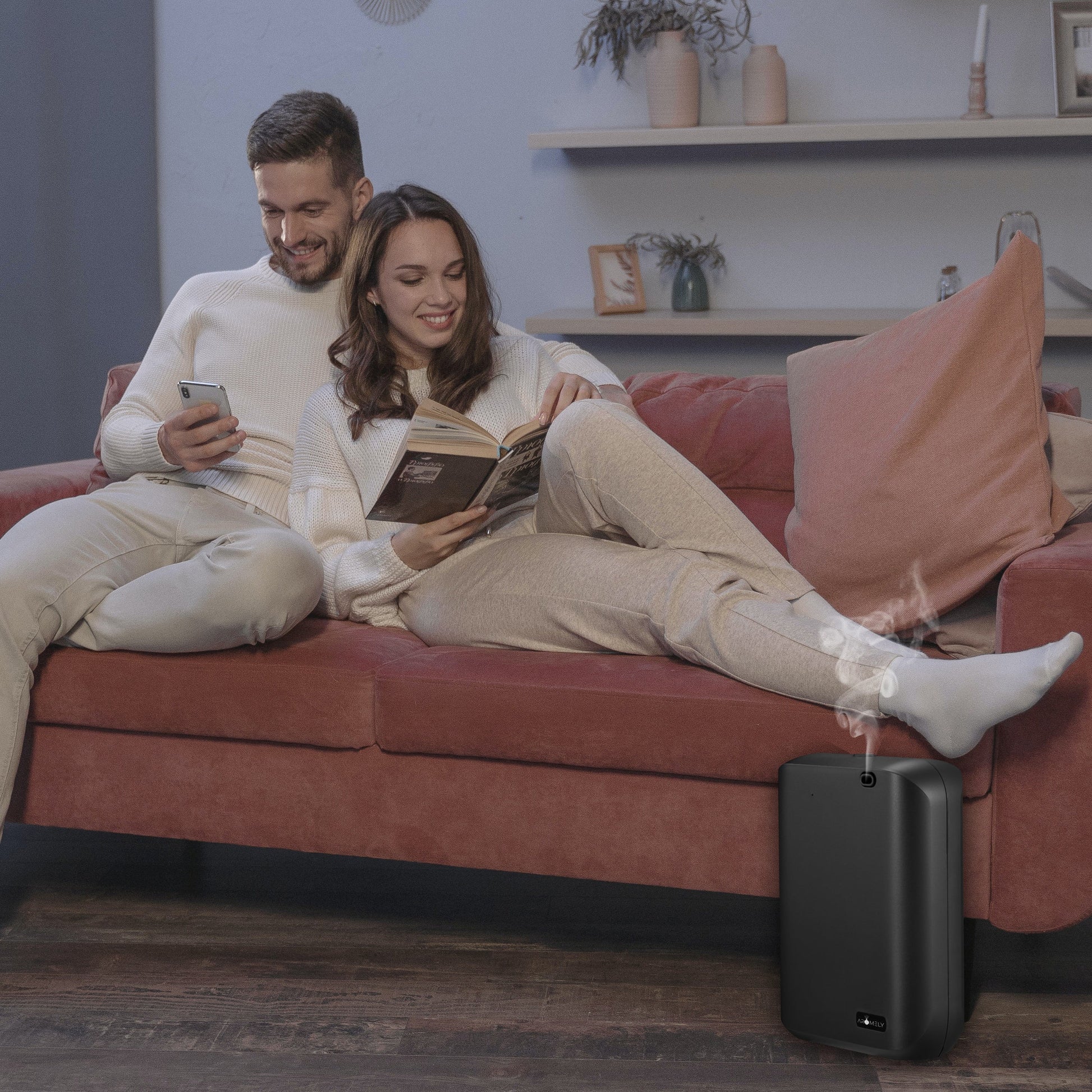 A man and woman unwind on a red sofa; the man checks his phone while the woman reads. Nearby, an AROMELY Smart Scent Diffuser fills the room with fragrance using cold air diffusion, perfect for spaces up to 1,000 SQFT.