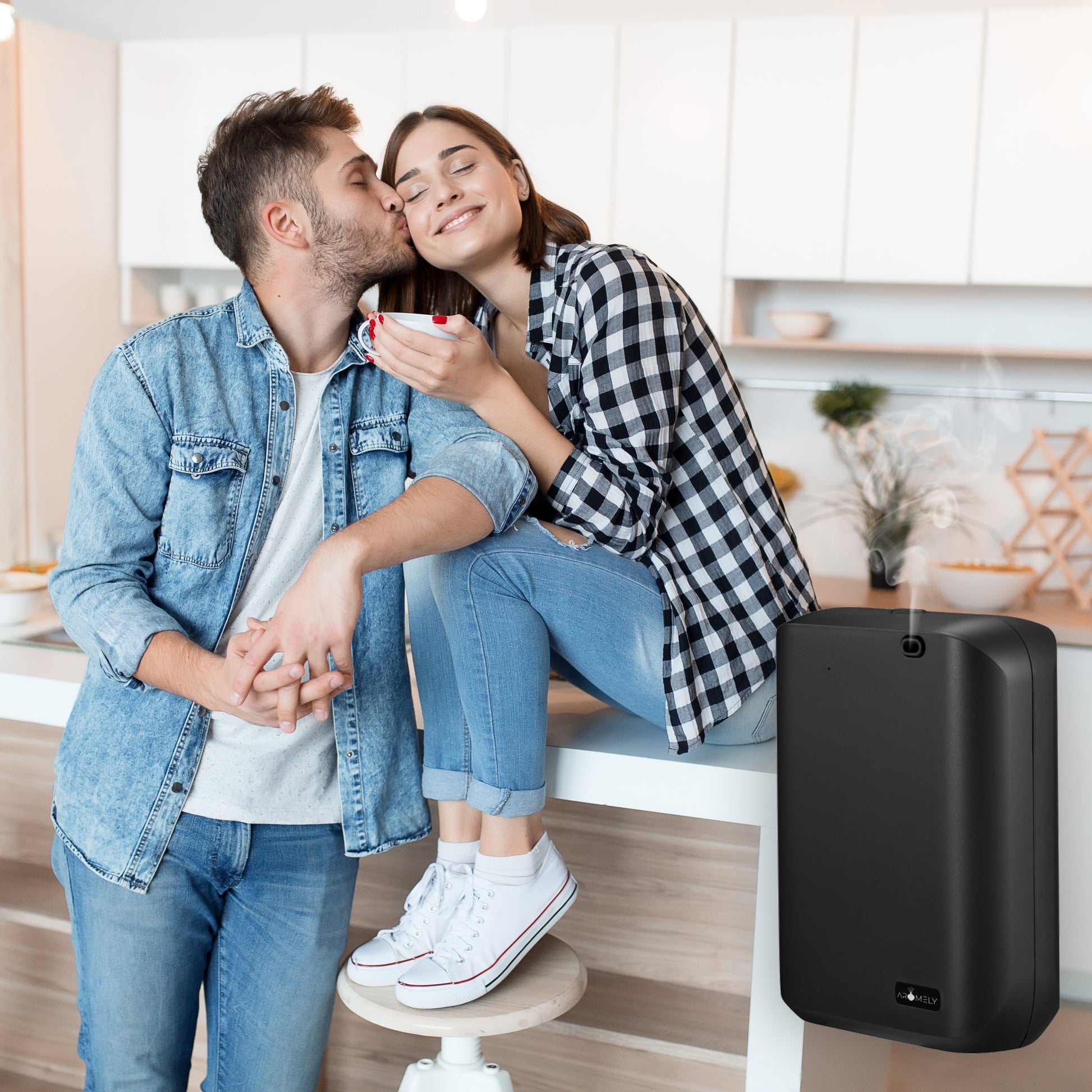 A man kisses a smiling woman on the cheek as she holds a mug in their bright kitchen, while an AROMELY Aromely Smart Scent Diffuser for Home fills the air with a gentle essential oil aroma.