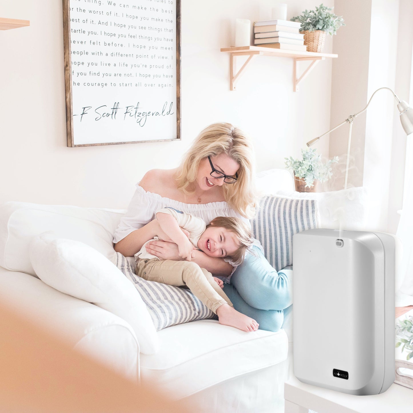A woman and child sit on a white couch in a bright living room, with an AROMELY Smart Scent Diffuser for Home, Office & SPA (up to 1,000 SQFT) using cold air diffusion in the foreground.