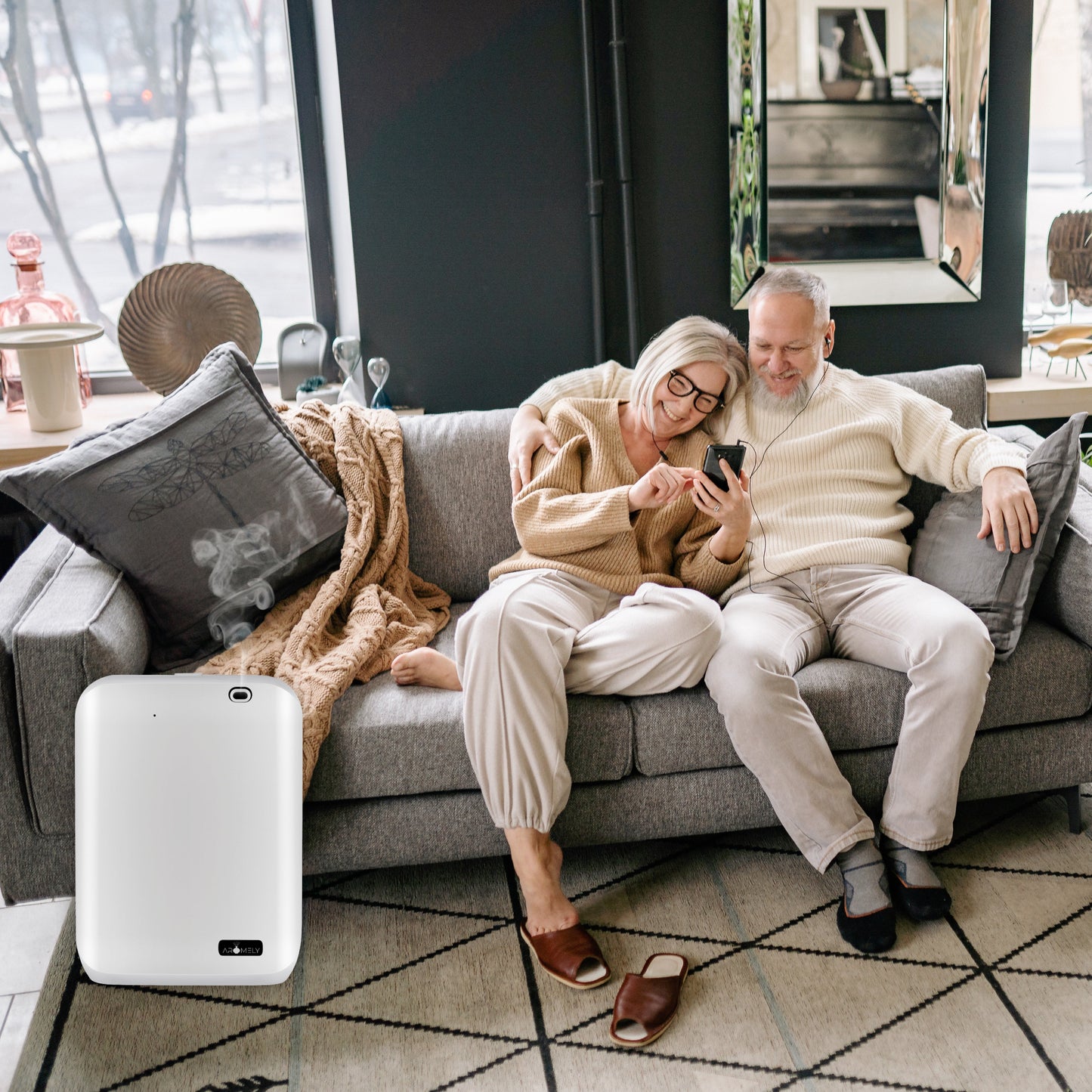 An older couple sits on a gray couch looking at a phone; nearby, an AROMELY Smart Scent Diffuser for Home, Office & SPA (covers up to 1,000 SQFT) rests on the floor by the sofa.