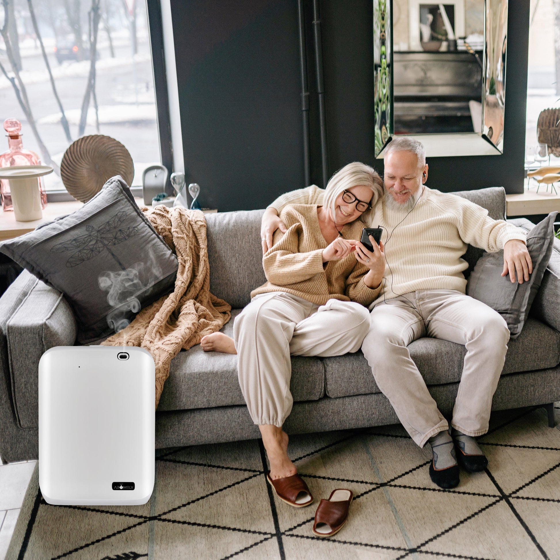 An older couple sits on a gray couch looking at a phone; nearby, an AROMELY Smart Scent Diffuser for Home, Office & SPA (covers up to 1,000 SQFT) rests on the floor by the sofa.