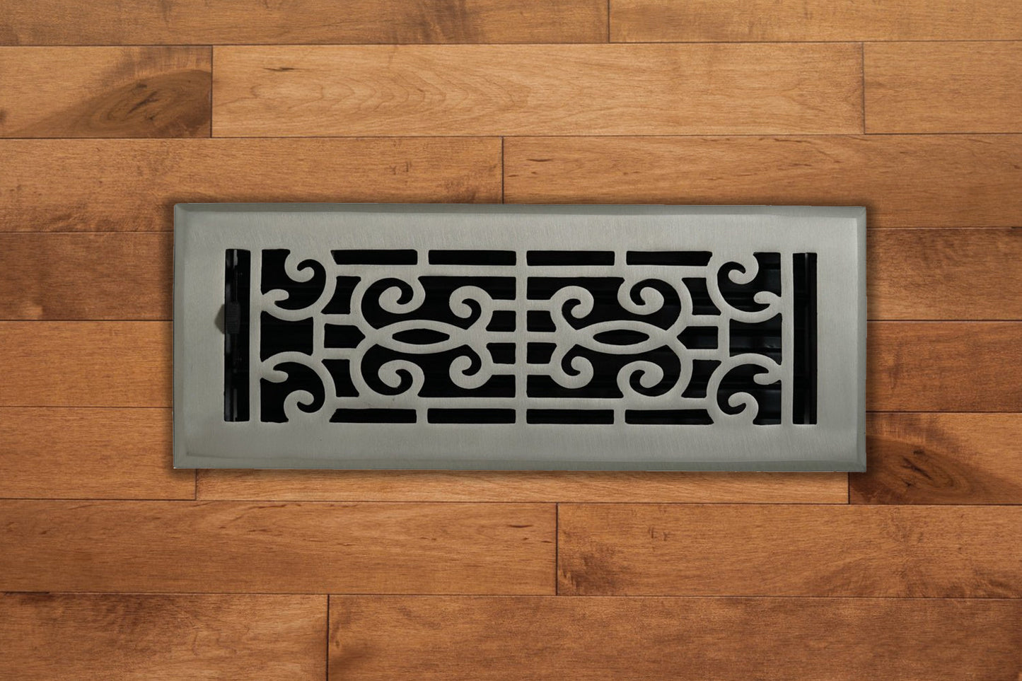 A Madelyn Carter Cast Brass Baroque Vent Cover in brushed nickel, featuring elegant scrollwork, is installed on a hardwood floor.