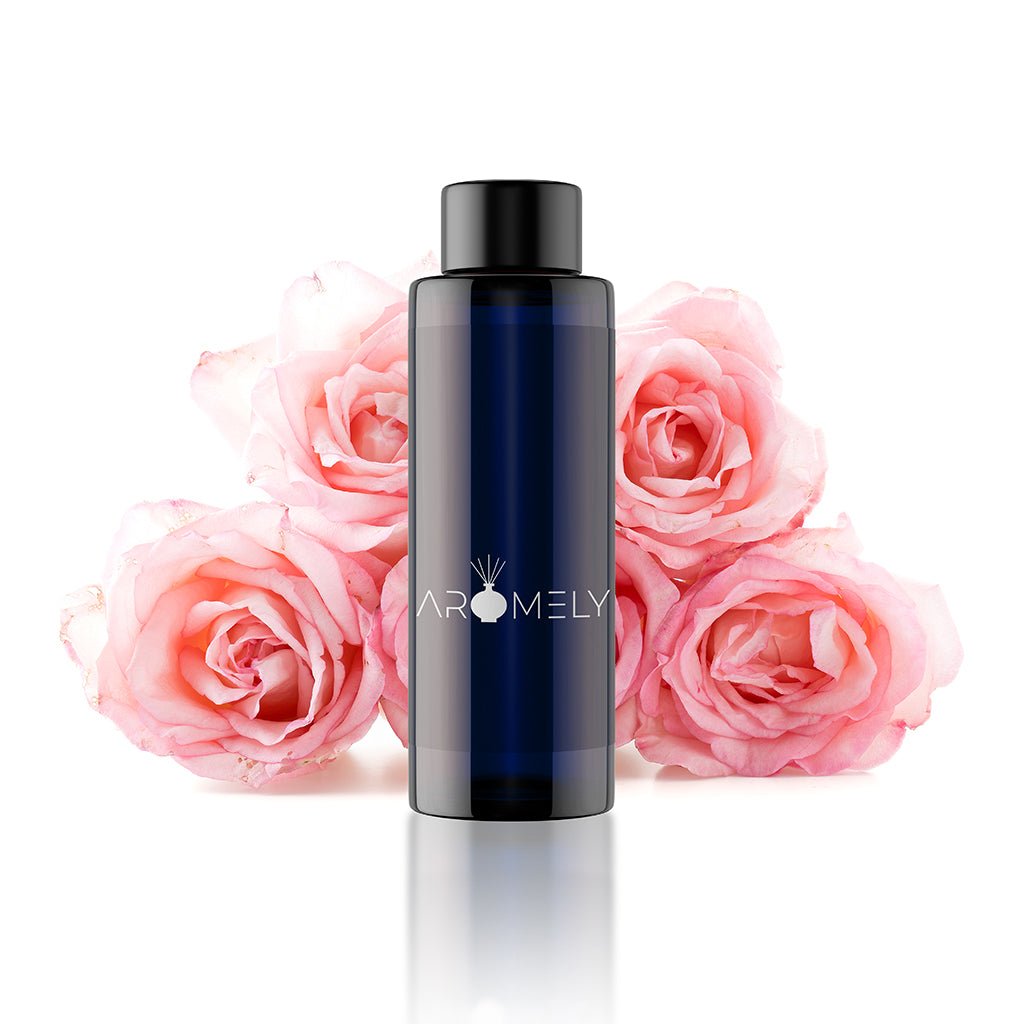 A dark blue AROMELY bottle labeled "Bloom Bliss - Inspired by: Fairmont® Hotels" is placed before four pink roses on a white background, capturing the allure of this romantic rose oud perfume.