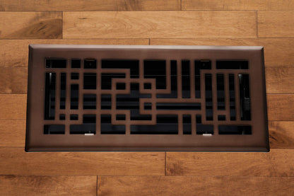 The Madelyn Carter Steel Capitman Art Deco Vent Cover in oil rubbed bronze, featuring a geometric pattern, is installed on a light brown wooden floor.