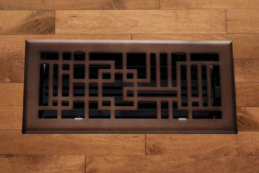 The Madelyn Carter Steel Capitman Art Deco Vent Cover in oil rubbed bronze, featuring a geometric pattern, is installed on a light brown wooden floor.