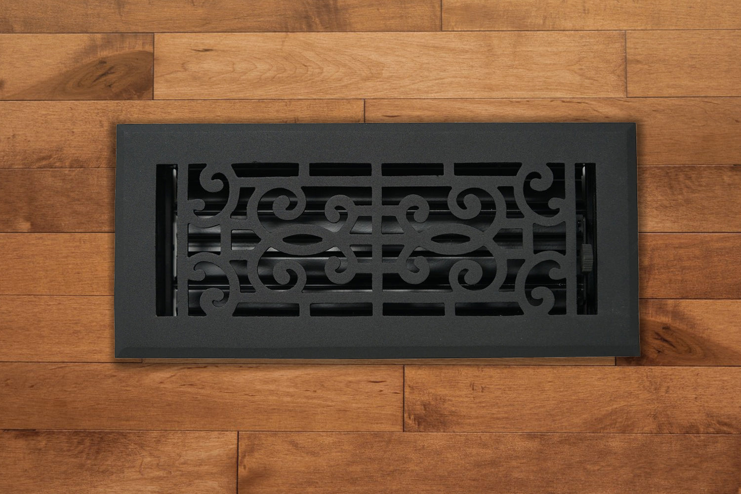 The Madelyn Carter Cast Aluminum Baroque Vent Cover in black, shown on a light brown wooden floor, adds elegance and airflow control with its scroll design and adjustable metal dampers.