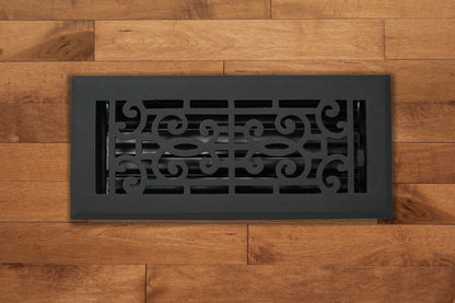 The Madelyn Carter Cast Aluminum Baroque Vent Cover in black, shown on a light brown wooden floor, adds elegance and airflow control with its scroll design and adjustable metal dampers.